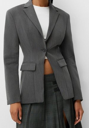 Woman wearing a fitted grey blazer with one button, over a white shirt, paired with a dark grey pleated skirt with a thigh slit.