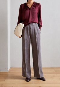 Burgunderfarbige Seidenbluse mit V-Ausschnitt und Puffärmeln, kombiniert mit lockeren, hellgrauen Leinenhosen. Das Modell hält eine cremefarbene Clutch.