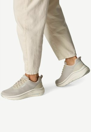 Person wearing beige mesh sneakers and light khaki pants, walking on a white background with one foot slightly raised.