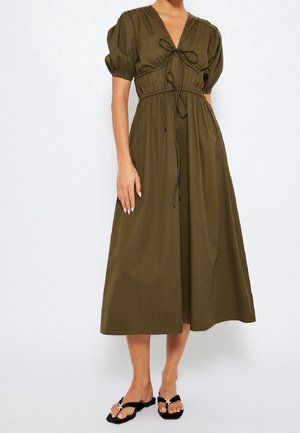 Mujer vistiendo un vestido midi verde oliva con mangas cortas abullonadas, detalles de lazo en el frente y sandalias planas negras con adornos de estrella de mar.