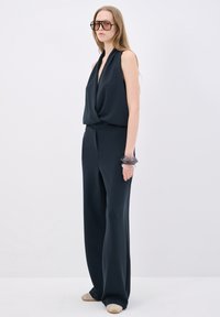 Marinblå ärmlös jumpsuit med djup V-halsringning och avslappnad passform. Har vida byxor och ett genomskinligt armband som accent på handleden.