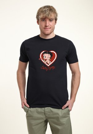 Ung mand iført sort t-shirt med rød hjerte og Betty Boop-figur grafik, kombineret med lysegrønne bukser, står med hænderne i lommerne.