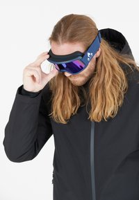 Hiihtolasit sinisellä hihnalla ja violetilla linssillä, joissa on pehmeä vaahtopehmuste. Käyttäjä säätää lasejaan mustan takin kanssa.