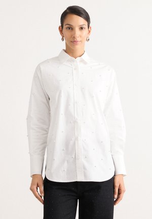Hvit skjorte med strukturert perlepynt, lange ermer og spiss krage. Brukt med mørke, pyntede jeans.