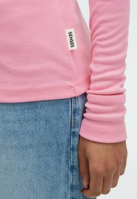 Helle rosa Langarmbluse aus weichem Baumwollstoff, mit einer genähten Saumkante und einem kleinen Etikett mit der Aufschrift "SENSES" an der Seite. Kombiniert mit Jeans.