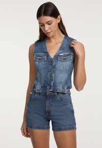 Jeans prsluk s V-dekolteom, prednjim gumbima i dva džepa na prsima. Nošen s usklađenim traper bermudama, oba u srednjem plavom pranju.