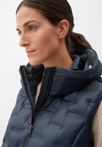Dunkelblauer Steppweste mit hohem Kragen, die über einem hellbeigen Oberteil getragen wird und ein gestepptes Design sowie einen Frontreißverschluss aufweist.