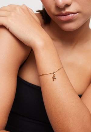 Femme portant un bracelet chaîne en or avec un petit pendentif en forme de croix à son poignet, habillée d'un haut noir sans manches.