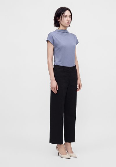 Femme debout portant un haut bleu clair à manches courtes, un pantalon noir à jambes larges et des escarpins beige pointus sur un fond uni.
