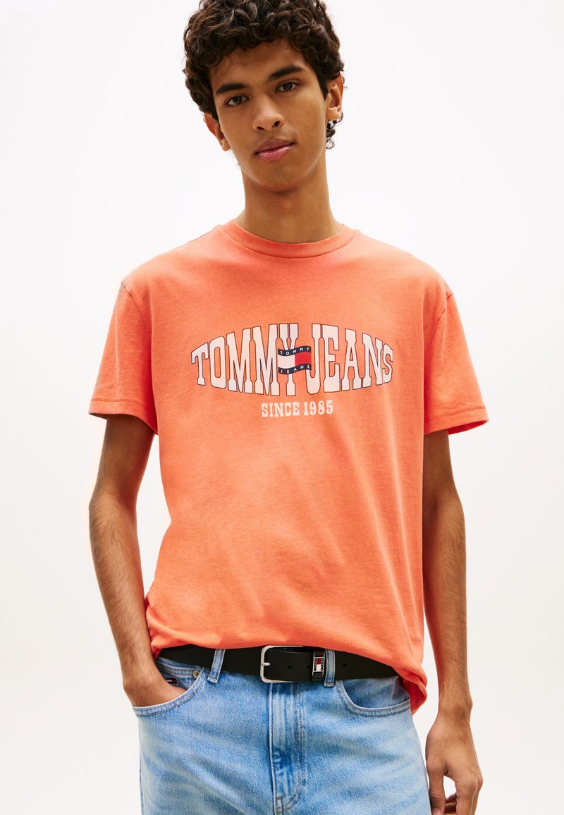 Νέος άνδρας που φοράει κοραλί μπλουζάκι Tommy Jeans και ανοιχτόχρωμο τζιν, με το ένα χέρι στην τσέπη, μπροστά σε απλό φόντο.