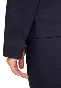 Marinblå kavajsärm med en vent, som visar upp en skräddarsydd design och slät textur. Handen placerad nära en söm på sidan av en åtsittande plagg.