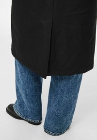 Giacca nera dal taglio dritto, abbinata a jeans blu a gamba larga con una texture sbiadita e cuciture a contrasto, completata da scarpe nere.