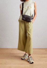 Person i ärmlös beige topp, ljusgröna vida byxor, beige och svarta sneakers, och en svart crossbody-väska står på träddäck.