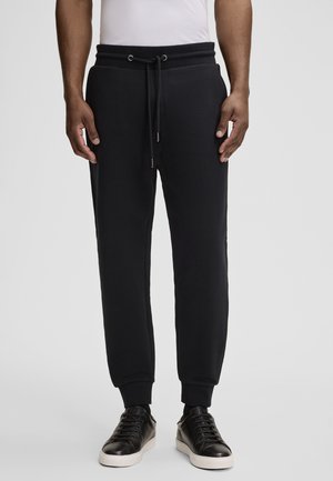 Schwarze Jogginghose aus weichem Material, mit einem Kordelzugbund, Seitentaschen und gerippten Bündchen. Kombiniert mit schwarzen Sneakern.