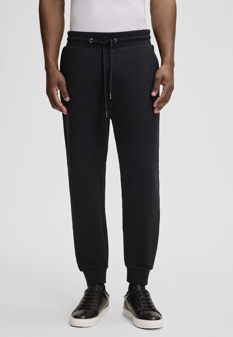 Schwarze Jogginghose aus weichem Material, mit einem Kordelzugbund, Seitentaschen und gerippten Bündchen. Kombiniert mit schwarzen Sneakern.