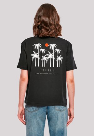 Person with shoulder-length brown hair wears black T-shirt with white palm trees and orange sun graphic, paired with blue jeans.