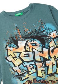 Tyrkysové bavlněné tričko s výrazným graffiti-style nápisem v žluté a černé barvě, doplněné uměleckými detaily městského panoramatu a obrázkem velryby.