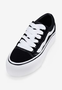 Zapatilla de ante negra con suela de goma blanca, con cordones blancos y detalles de franjas laterales. Punta redondeada con acentos de costura negra.