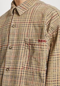 Plaid button-up shirt in beige with blue, red, and green patterns. Features a chest pocket and embroidered "BDG" logo. Textured fabric.
