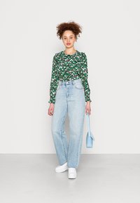Blusa verde floral com decote franzido, calças de ganga de cintura alta azul claro, ténis brancos e uma pequena mala azul.
