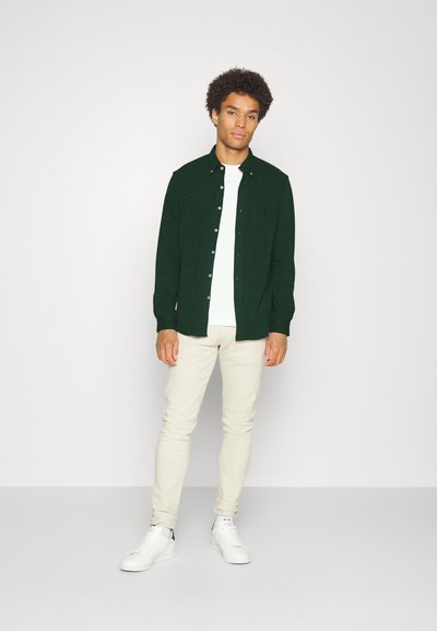 Groene overhemd met knopen over een wit T-shirt, gecombineerd met lichtbeige broek en witte sneakers. Eenvoudig ontwerp zonder noemenswaardige patronen.