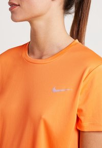 Oranžové sportovní tričko z hladkého, lehkého materiálu, s malým fialovým logem Nike na hrudi a klasickým kulatým výstřihem.
