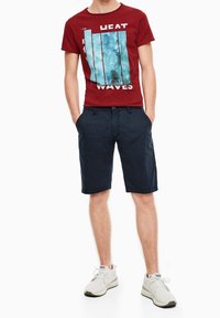 Muž oblečený v červeném tričku s grafikou, námořnických modrých šortkách a bílých teniskách, stojící s rukama v kapsách na bílém pozadí.