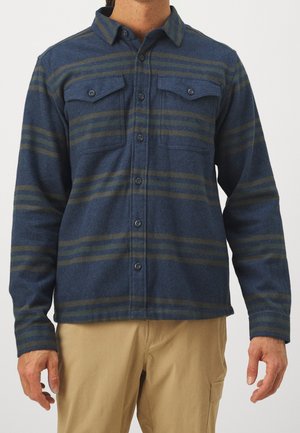Homme portant une chemise rayée bleue et verte à boutons avec deux poches poitrine et un pantalon beige sur un fond uni.