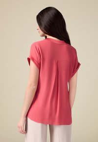 Blusa corallo a maniche corte con vestibilità rilassata, dettaglio a pieghe sul retro e orlo leggermente arrotondato, realizzata in tessuto leggero.