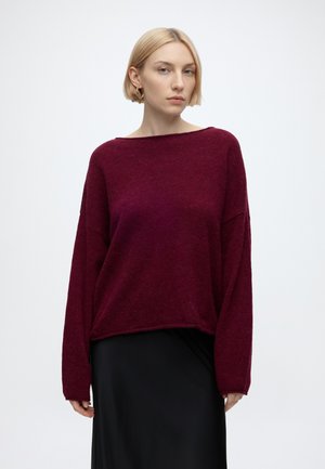 Femme aux cheveux blonds courts portant de grandes boucles d'oreilles créoles, un pull bordeaux ample et une jupe noire sur un fond blanc uni.