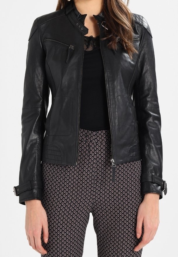 Femme portant une veste en cuir noire cintrée avec fermeture éclair et détails de boucles sur un haut noir et un pantalon à motifs.
