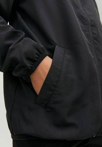 Černá lehká bunda s elastickými manžetami a bočními kapsami, s jemnou texturou a minimalistickými designovými prvky.