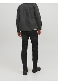 Tmavě šedá dlouhá košile s límečkem na knoflíky, v kombinaci s černými slim-fit džíny a tmavými botami, pohled zezadu.