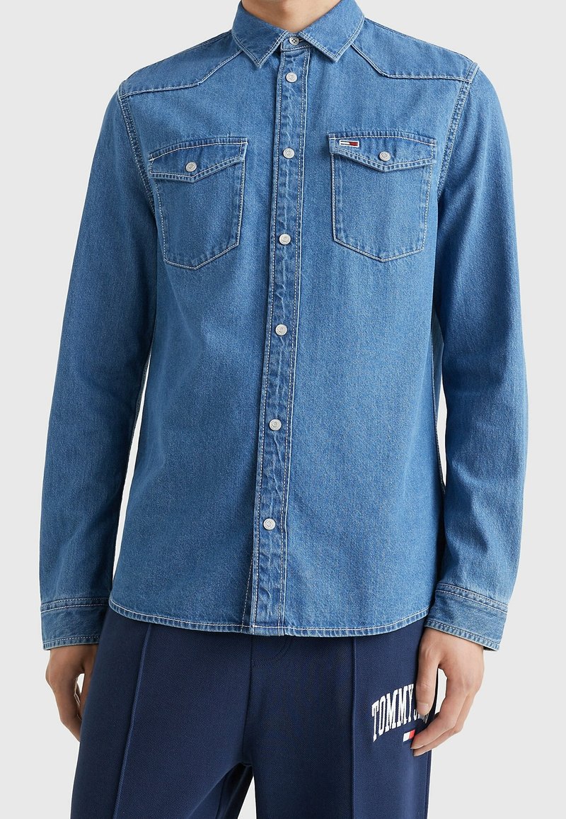 Blå denimskjorta med en figurnära design, spetsig krage och två bröstfickor med knappar. Inkluderar kontrastsömmar och en liten logotyp.