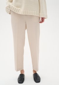 Beige skræddersyede bukser med lige pasform, der har en glat tekstur og diskrete folder, samt sorte slip-on sko.