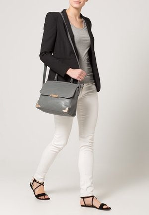 Femme portant un blazer noir, un haut gris, un pantalon blanc, des sandales noires, et un sac à main en cuir gris avec des accents dorés porté à l'épaule.