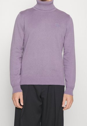 Personne portant un pull à col roulé lavande à manches longues et un pantalon noir ample, debout devant un fond clair uni.