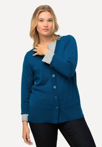 Tealgul cardigan med fem knappar, ribbade ärmslut och en strukturerad yta, lagd över en grå topp. Bärs med mörka jeans.