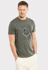 T-shirt en coton vert olive à manches courtes, col rond, avec un imprimé graphique en marine et crème, présentant des rayures horizontales et un design vintage.