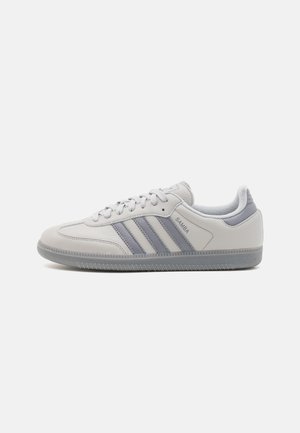 Biały sneaker Adidas Samba z szarymi paskami, sznurówkami i teksturowaną podeszwą, pokazany na boku na białym tle.
