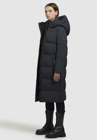 Mørk puffjakke med hætte, med et quiltet design, frontlynlås og sidelommer. Båret over sorte leggings og chunky sorte støvler.