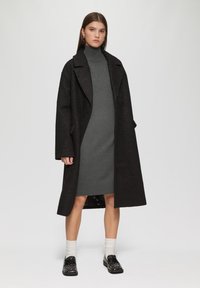 Manteau noir texturé, robe à col roulé grise, chaussettes crème et chaussures noires à clous. Le mannequin se tient sur un fond uni.