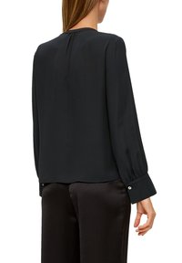 Schwarze Bluse mit langen Ärmeln aus glattem Stoff, mit einem runden Ausschnitt und geknöpften Manschetten. Der Rücken ist etwas länger und hat einen lässigen Schnitt.