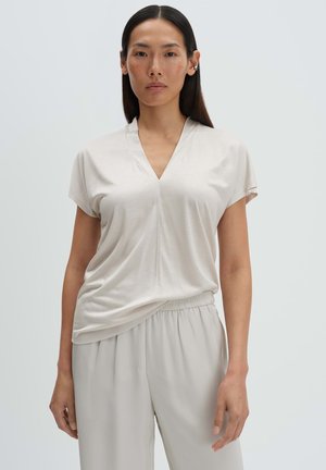 Vrouw met lang donker haar, gekleed in een licht beige top met V-hals en bijpassende losse broek, staand tegen een effen achtergrond.