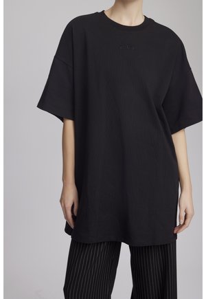 Personne portant un T-shirt noir oversize à manches courtes et un pantalon noir à fines rayures sur un fond uni.