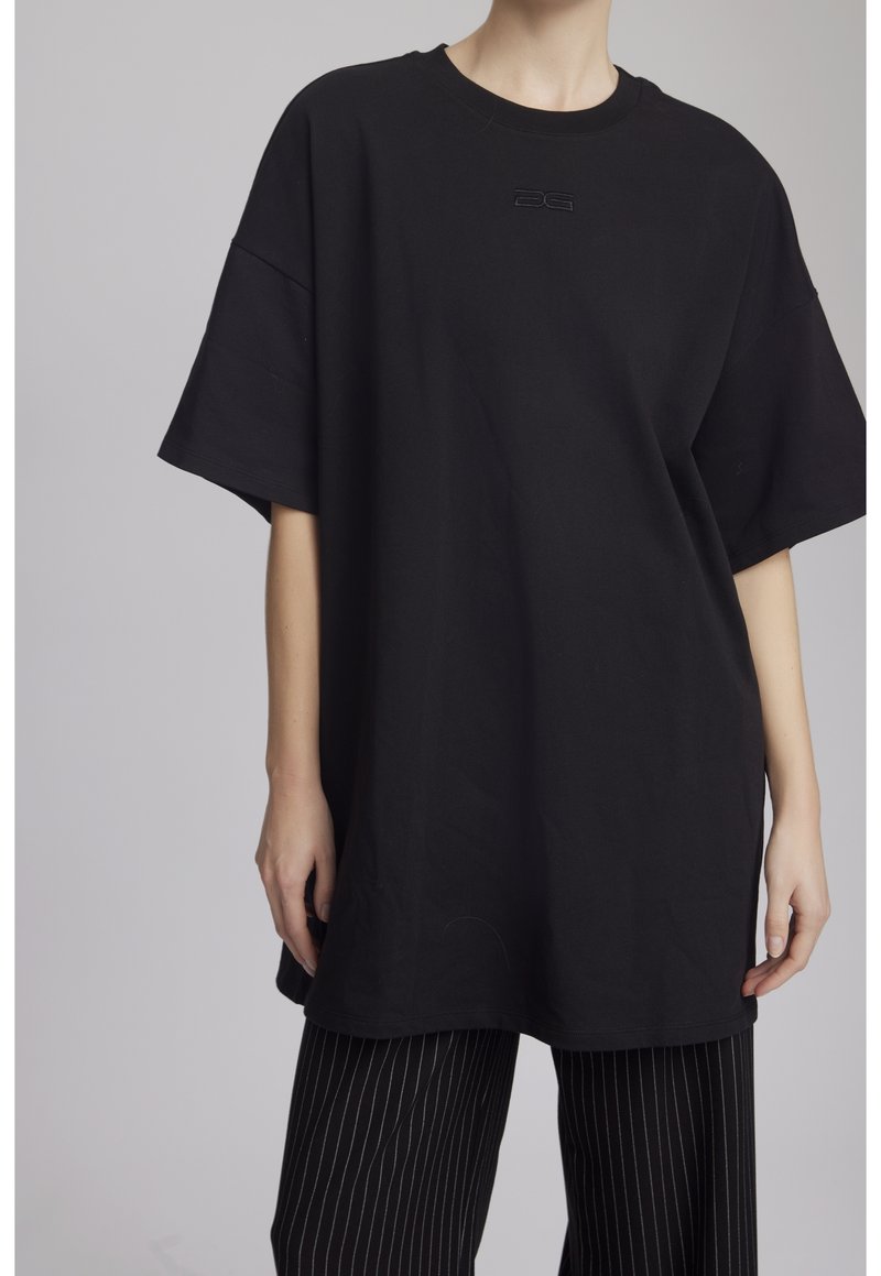 Personne portant un T-shirt noir oversize à manches courtes et un pantalon noir à fines rayures sur un fond uni.