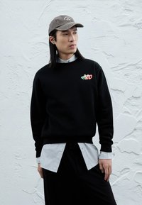 Schwarzer Pullover mit gesticktem Logo, darüber getragen ein gestreiftes Hemd mit Knöpfen. Eine graue Mütze vervollständigt das Outfit, vor einem strukturierten weißen Hintergrund.