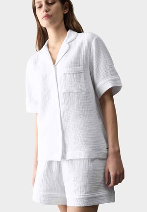 Kvinde iført hvid tekstureret kortærmet skjorte med brystlomme og matchende shorts mod en ensfarvet baggrund.