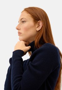 Maglione lavorato a maglia di colore blu navy, con colletto alto e maniche lunghe, che presenta una consistenza morbida e un design aderente, indossato da una persona con capelli rossi.
