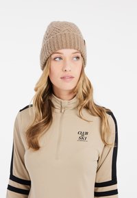 Beige gestrickte Mütze mit einem strukturierten Kabelmustern und einem gefalteten Rippbündchen. Das Model trägt ein beige zipfähiges Oberteil mit schwarzen Akzenten und Logo.